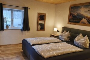 Ein Schlafzimmer mit Bett, Kissen, Nachttisch, Lampe, einem Fenster mit Vorhängen, einem Spiegel und einem Bild an der Wand.