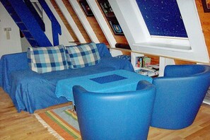 Ein Zimmer mit einem blauen Sofa, zwei blauen Sesseln, einem Fenster mit Blick auf einen Sternenhimmel und einer schrägen Decke.
