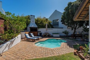 Un patio trasero con piscina, sillas de descanso, zona de barbacoa y una casa blanca con techo de paja.