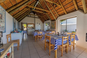 A dining area with striped tablecloths, wooden chairs, a kitchen in the background, and a thatched roof.