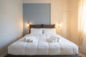 A neatly made bed with white linens and rolled towels, two wall-mounted light fixtures, and a plain wall with a door. - Terrace Penthouse Gargasole SX – Elegant 7th-Floor Retreat with Terrace in Bari (Bari)
