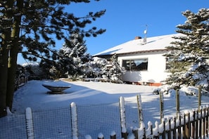 Ein mit Schnee bedecktes Haus mit einem eingezäunten Garten, einer verschneiten Gartendekoration und blauem, wolkenlosem Himmel.