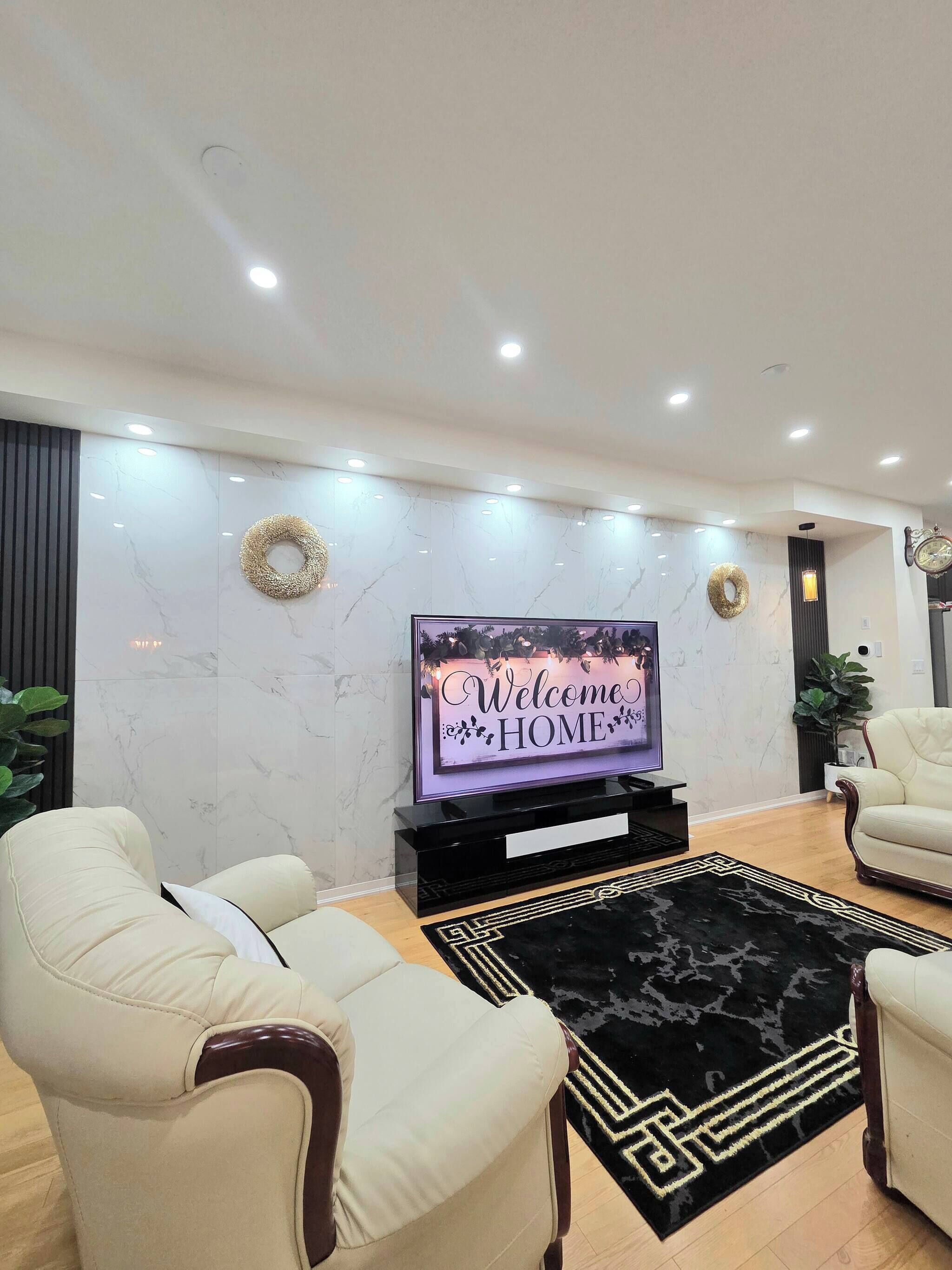 A modern living room with a flat-screen TV, two cream-colored armchairs, a black and gold patterned rug, and a marble wall with recessed lighting.