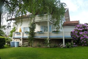 Ein zweigeschossiges Haus mit einem Balkon, umgeben von Grünpflanzen und Blumen.