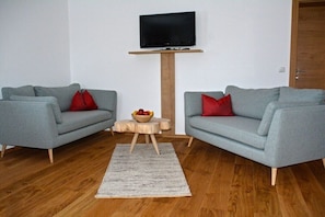 Ein Wohnzimmer mit zwei grauen Sofas, einem Holztisch, einem an einer Holzkonstruktion befestigten Flachbildfernseher und Holzboden.