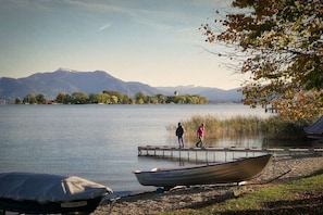 Ein See mit einem Boot, das an der Ufer angelegt ist, eine Person, die am Steg entlanggeht, und Berge im Hintergrund.