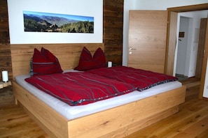 Ein hölzernes Bett mit roter Decke und Kissen, ein Holz-Kopfstück, ein gerahmtes Bild an der Wand und eine Holztür.