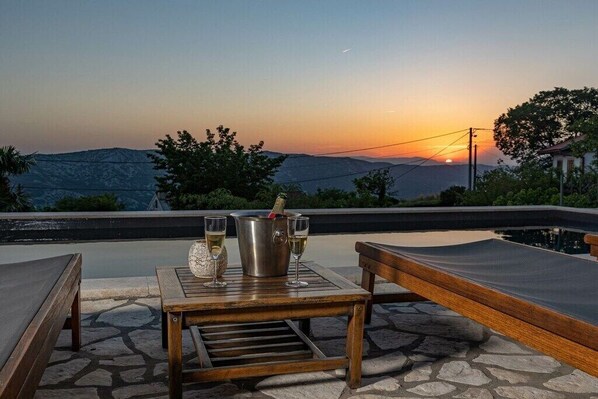 Une vue sur le coucher de soleil depuis une terrasse, avec une table, deux fauteuils et une bouteille de champagne.