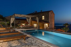 Un espace aménagé au bord d’une piscine en pierre, avec des chaises longues et un coin salon couvert offrant une vue sur le paysage.