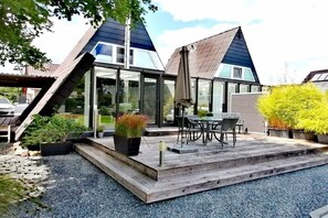Ein modernes Haus mit geneigtem Dach, großen Fenstern und einer Holzterrasse mit Gartenmöbeln.