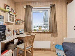 Ein Zimmer mit Holzeinrichtung, einem fenster mit Vorhängen, einem Schreibtisch mit diversen Gegenständen, einem Sessel, einem Heizkörper und einem Bett mit blauer Matratze.