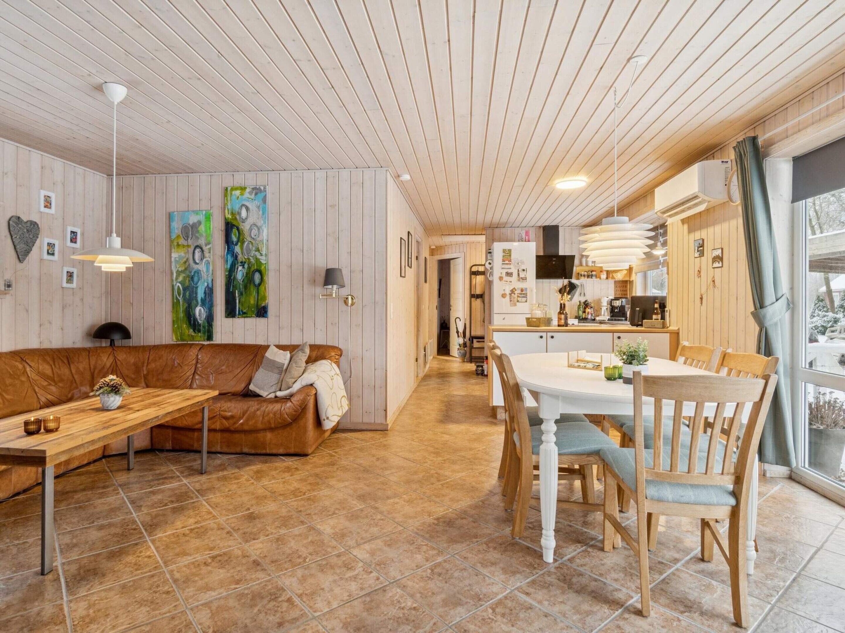 A cozy living area with a sofa, dining table, and chairs, wooden walls, and a kitchen in the background.