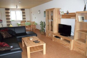 Ein Wohnzimmer mit schwarzem Sofa, Holztisch, TV-Standfuß und einer Essecke mit grünem Tisch und Stühlen.