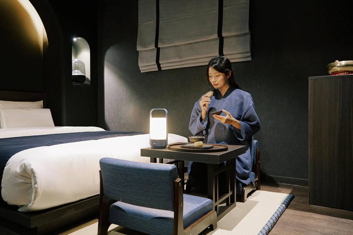 Una habitación de hotel con una cama, una mesa con tetera y tazas, una silla y una mesita de noche con lámpara.