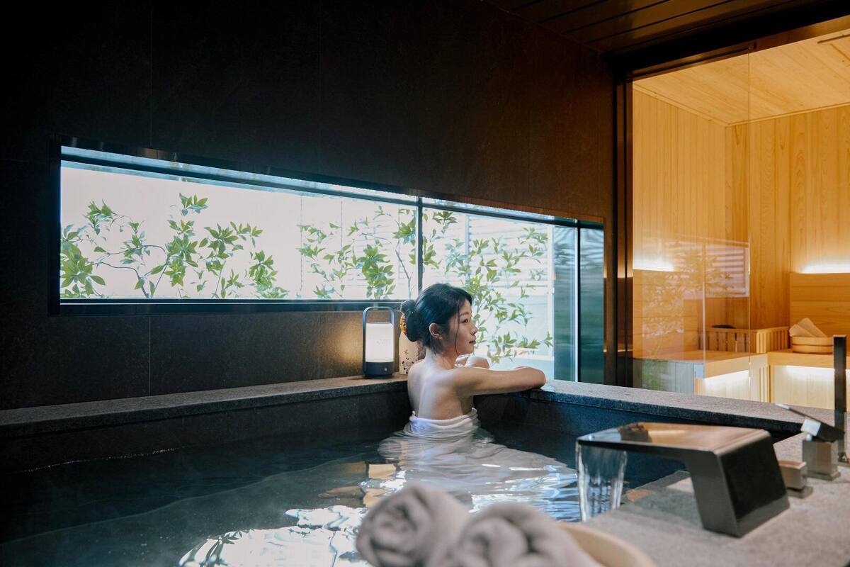 Una persona en un jacuzzi con vista a un cuarto con paredes de madera y una ventana con plantas.