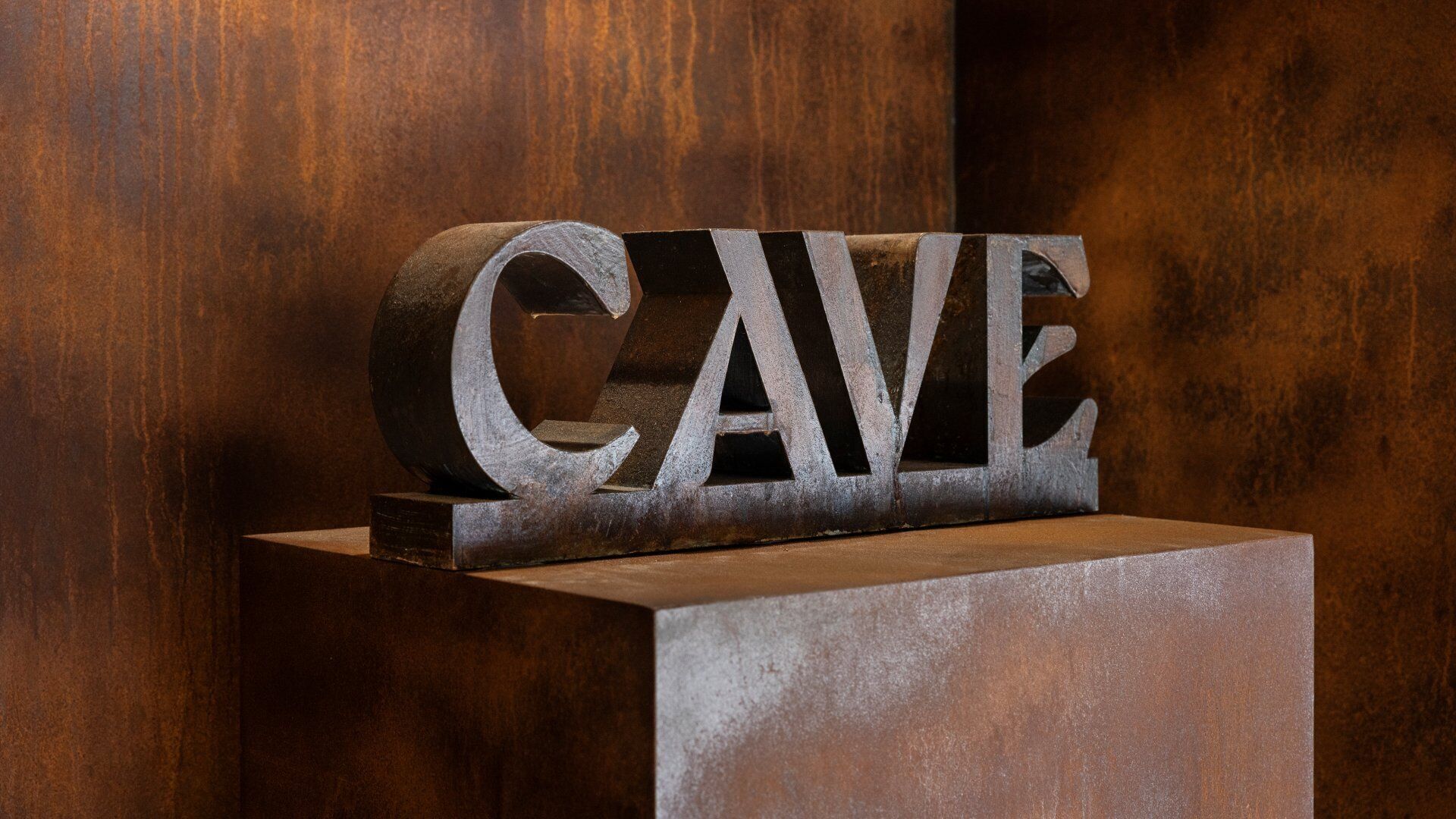 Una escultura de metal con la palabra 'CAVE' sobre un pedestal, frente a un fondo de madera.