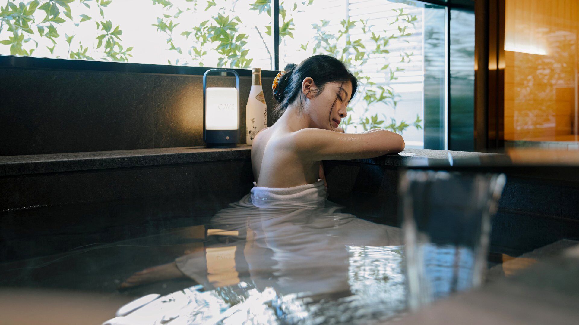 Una persona en un jacuzzi con una toalla alrededor del cuerpo, una botella y un pequeño dispositivo en un estante.