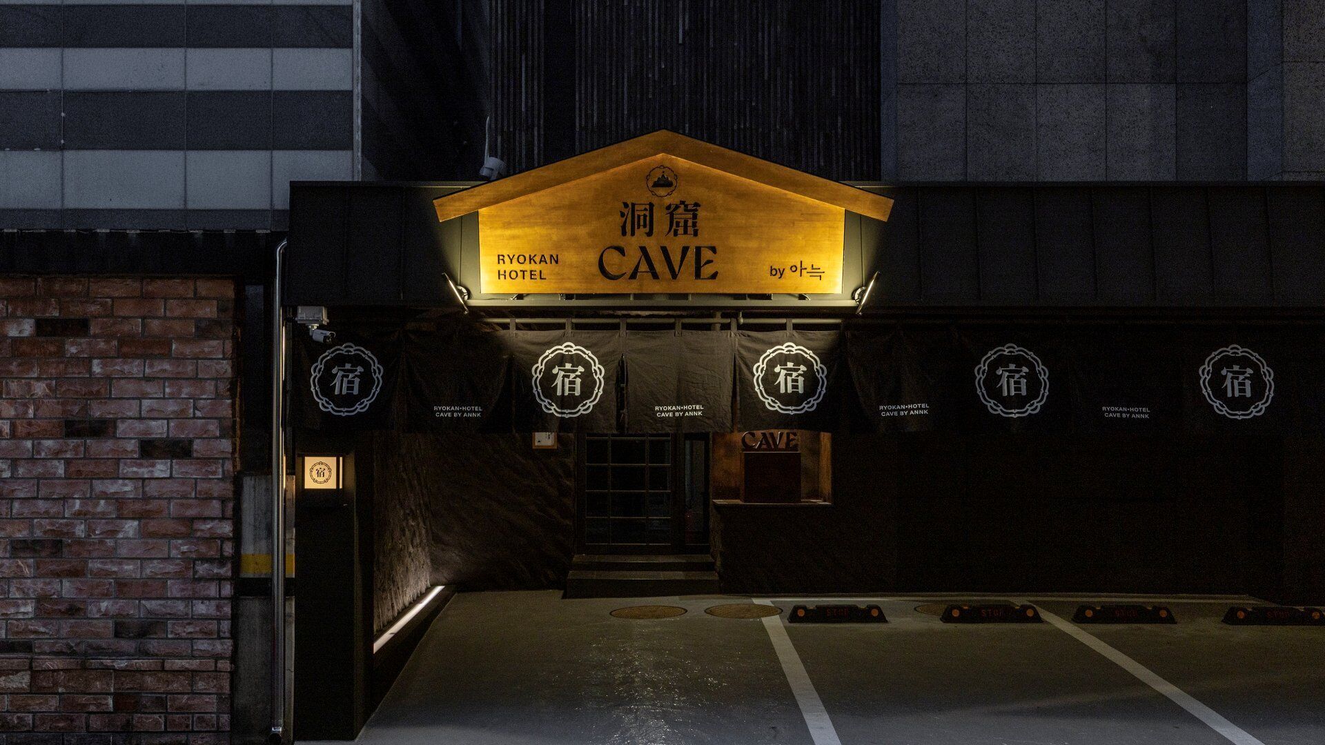Entrada al Hotel Ryokan Cave con diseño japonés tradicional, un letrero de madera y una pared de ladrillo.
