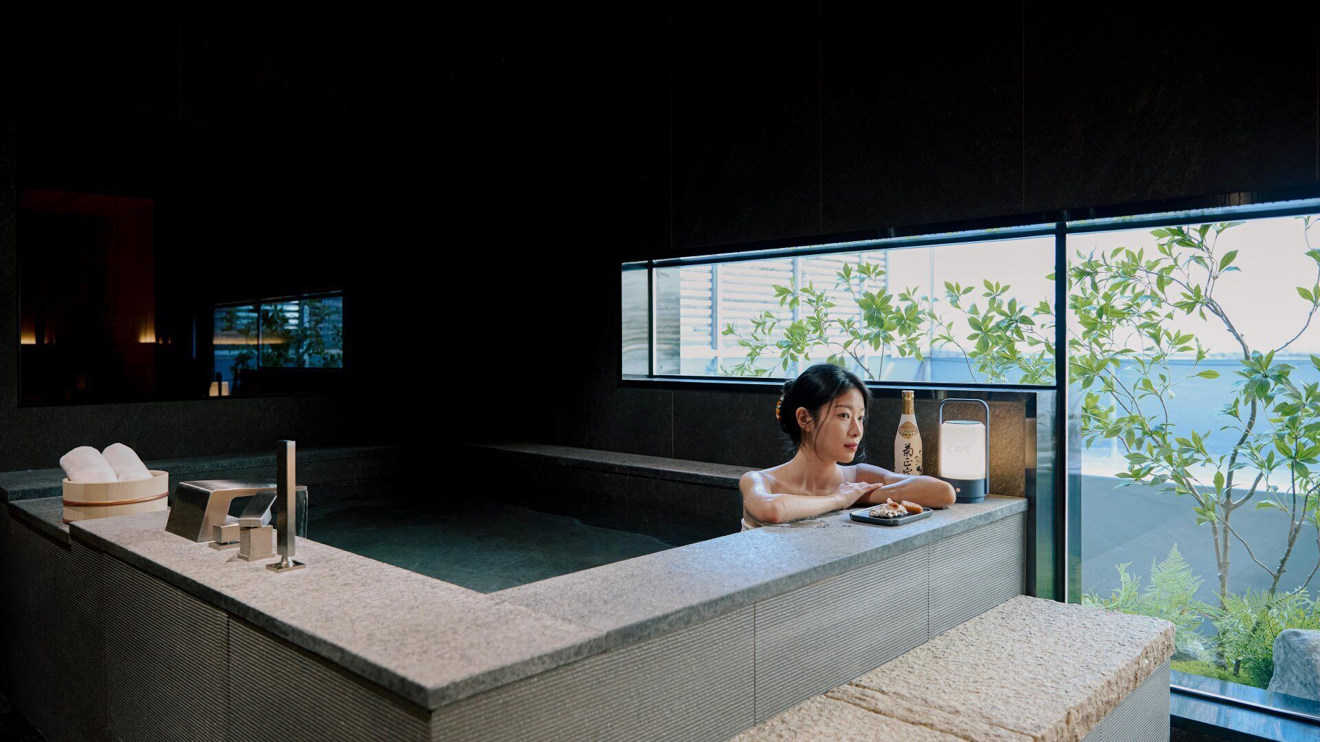 Un espace spa moderne comprenant une grande baignoire, un récipient en bois rempli de serviettes et une bouteille de saké.