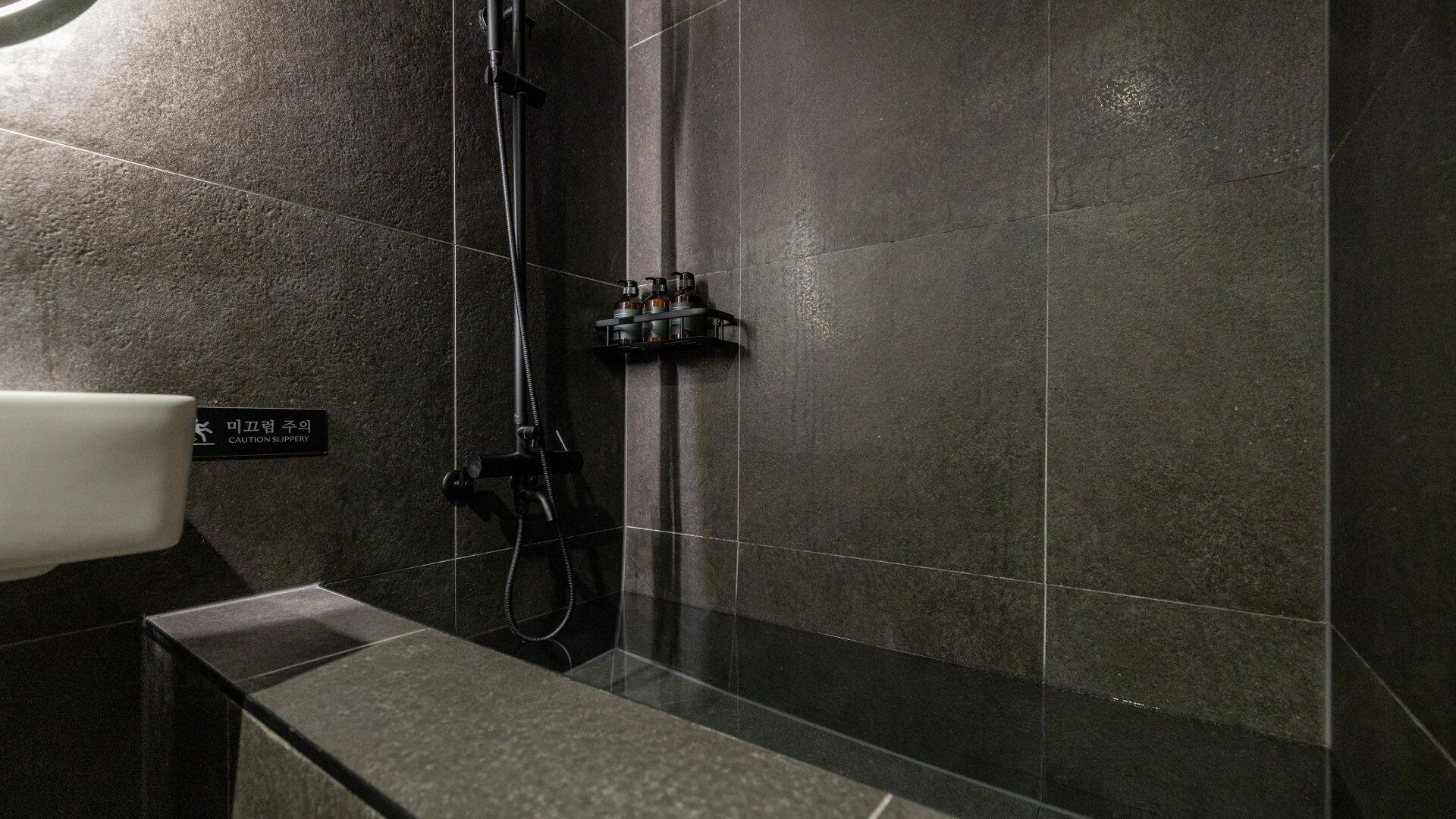 Une salle de bain moderne avec des carreaux foncés, un espace douche et un panneau d’avertissement indiquant de porter des chaussons antidérapants.