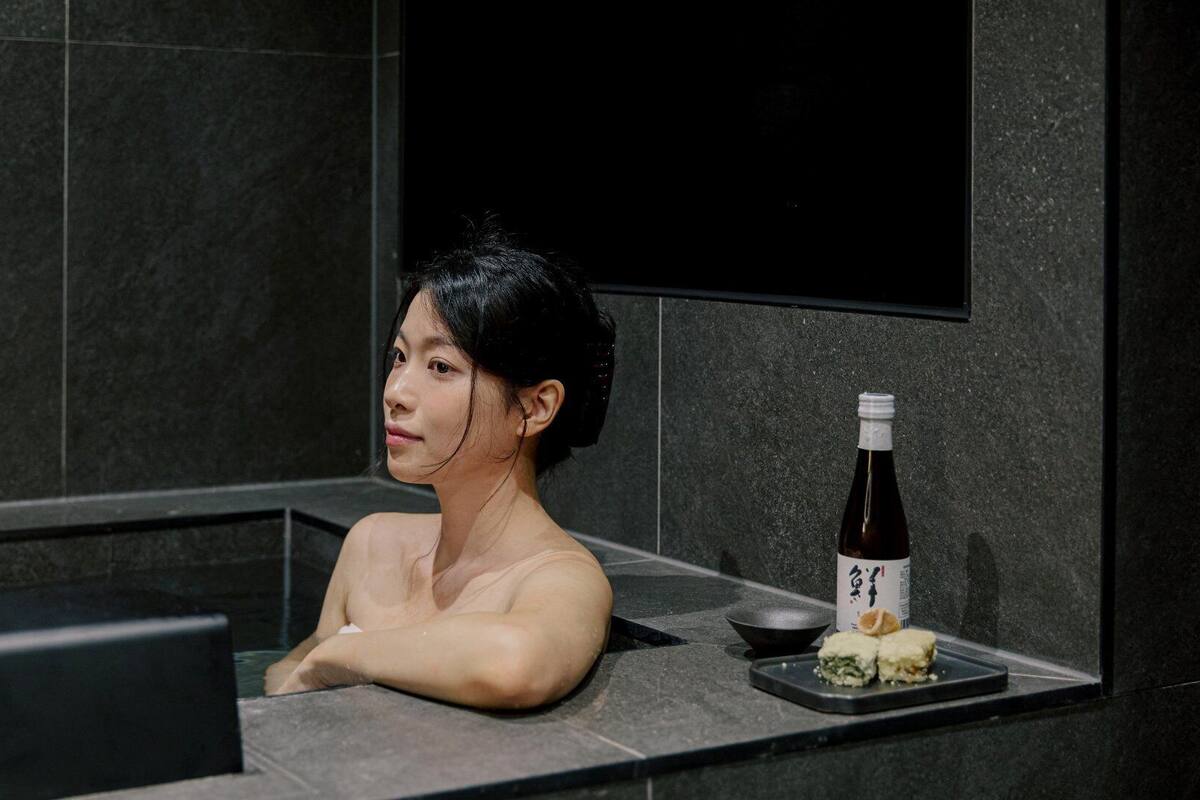 Una persona en una bañera con una botella de sake y un plato de comida a un lado.