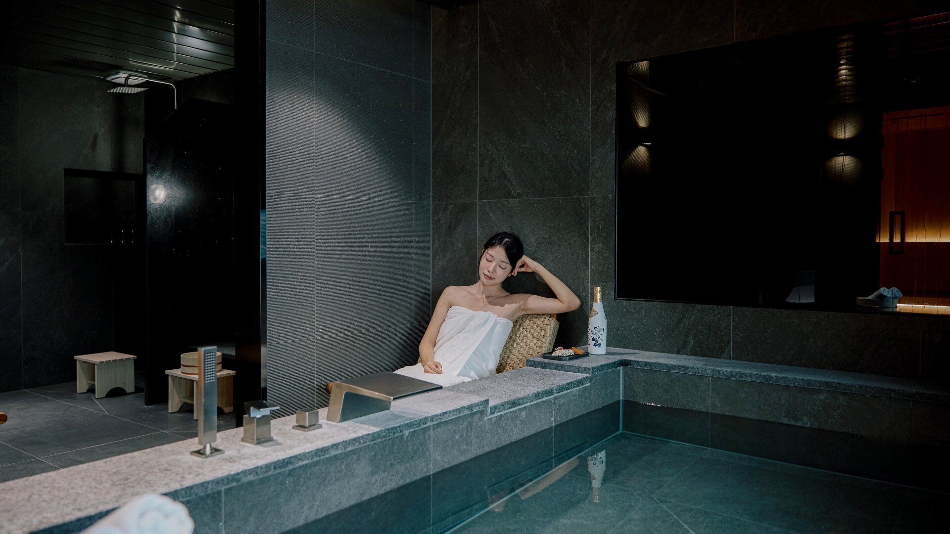 A person in a bathtub with a towel, a bottle, and a remote control on the counter.