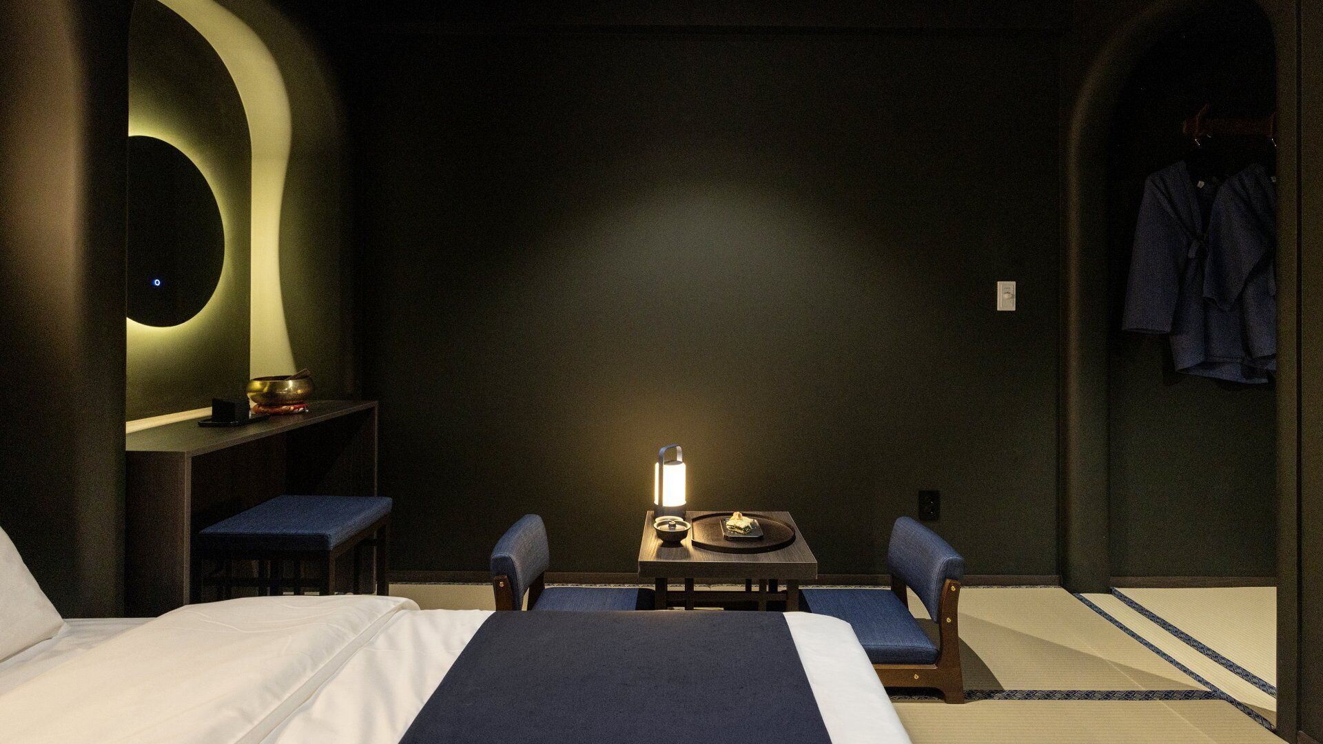 A hotel room with a bed, two chairs, a small table with a lamp, and a wall-mounted shelf with a bowl and a phone.