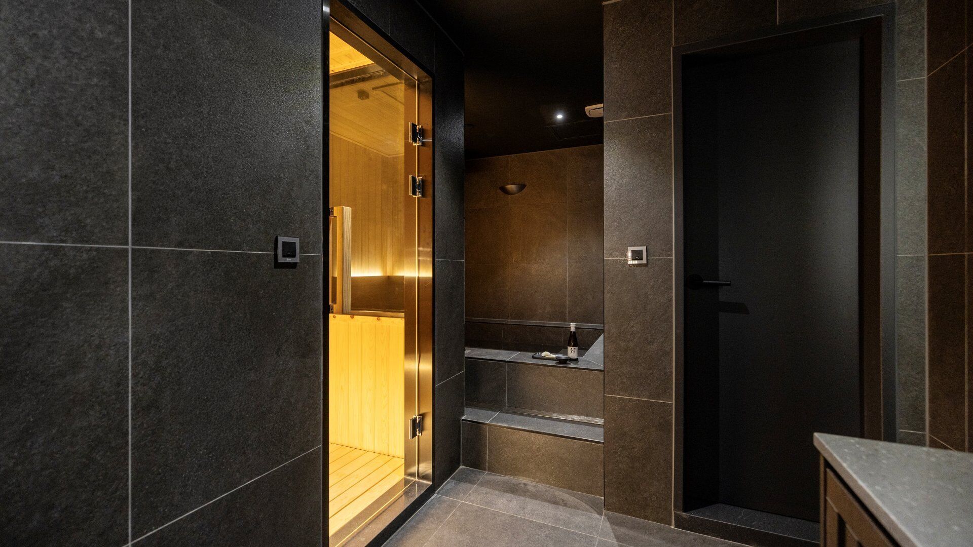 A modern bathroom with dark tiles, a wooden vanity, and a glass shower enclosure.