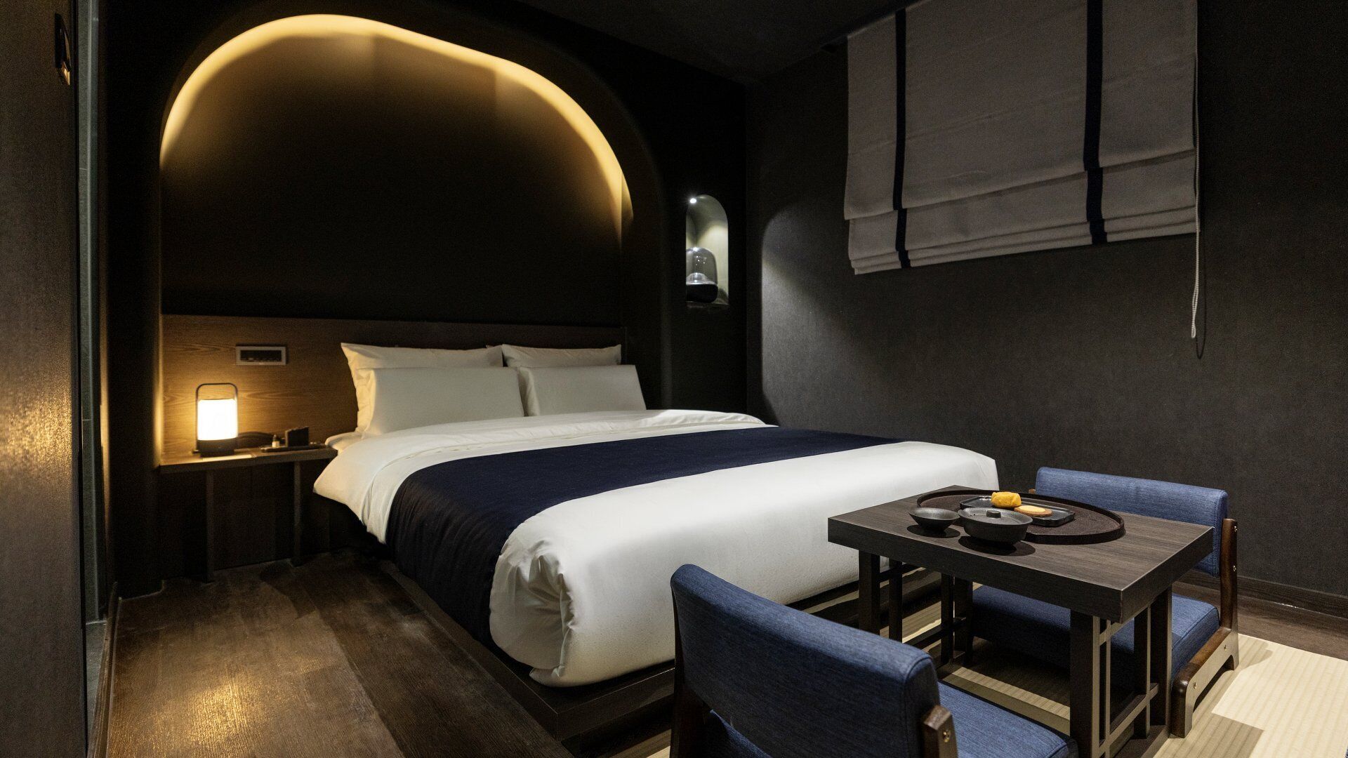 A hotel room with a bed, bedside table, lamp, and a small table with a chair.