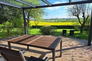 A covered patio with a wooden table and chairs, surrounded by a brick floor and a view of a green landscape with trees and fields. - Unique landscape view (Stoltebüll)