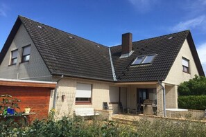 Ein zweistöckiges Haus mit geneigtem Dach, einer Holztür zur Garage und einer überdachten Terrasse mit Sitzgelegenheiten.