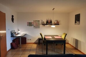 Ein Essbereich mit Tisch und Stühlen, ein Regal mit Flaschen, ein Schreibtisch mit Lampe und eine Wanduhr.