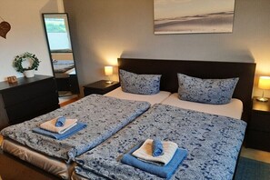 Ein Doppelbett mit gemusterten Bettbezügen, ein Nachttisch mit Lampe, ein Spiegel, ein an der Wand befestigtes Kunstwerk und ein kleiner Topfpflanze.