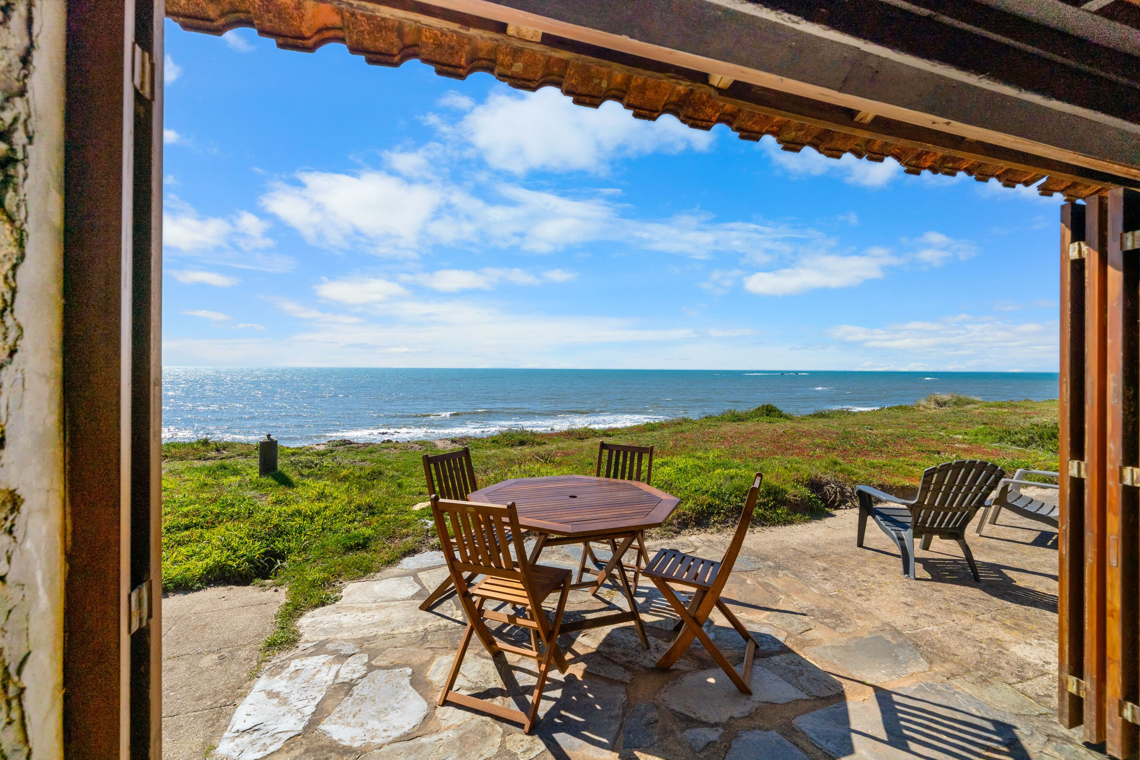 Eine Terrasse im Freien mit Tisch und Stühlen, mit Blick auf einen Strand, das Meer und den Himmel.