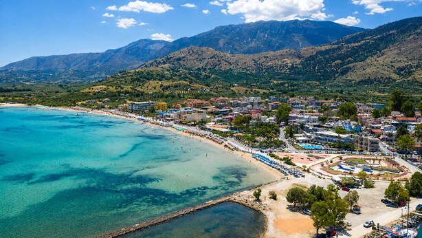 Uma vila costeira com praia, águas cristalinas e montanhas ao fundo.