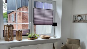 Une pièce comprenant un lit, une chaise, un radiateur, une fenêtre avec des stores, un bol en bois et une petite table avec des verres.