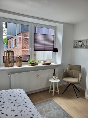 Ein Zimmer mit einem Bett, einem Sessel, einem Heizkörper, einem Fenster mit Jalousien, einer Holzschüssel und einem kleinen Tisch mit Gläsern.