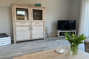 Ein Wohnzimmer mit einem Holzschrank, einem Fernseher auf einem Schrank, einer Glaskaraffe mit Blumen und einem Holztisch.