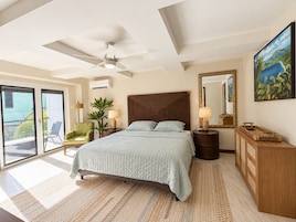 Un dormitorio con cama, ventilador de techo, mesita de noche, espejo, cómoda, silla y balcón con vistas.