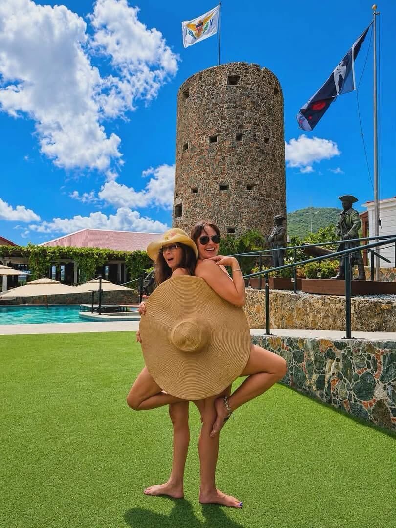 A person carrying another on their back near a pool with a stone tower in the background.