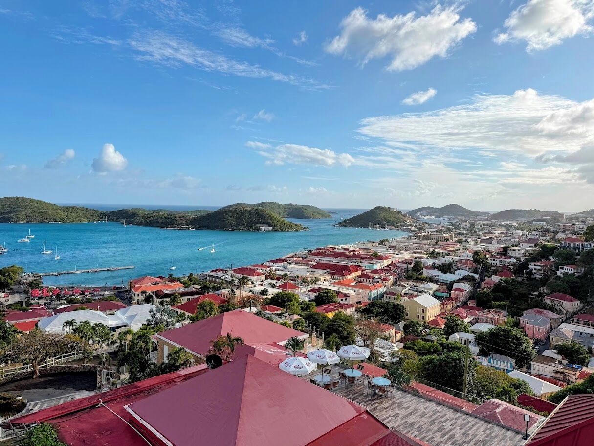 A coastal town with red-roofed buildings, a marina with boats, and a view of the sea and hills.