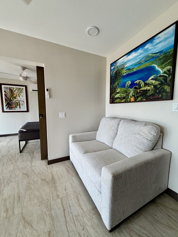 A living room with a grey sofa, a painting of a tropical landscape, a ceiling fan, and a small table with a chair.