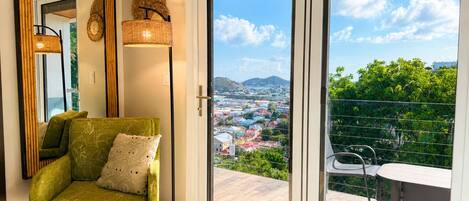 A room with a green sofa, a large mirror, a floor lamp, and a sliding glass door leading to a balcony with a view of a cityscape.