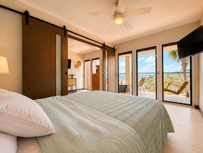 Un dormitorio con cama, ventilador de techo y vista a un balcón con silla y una palmera.