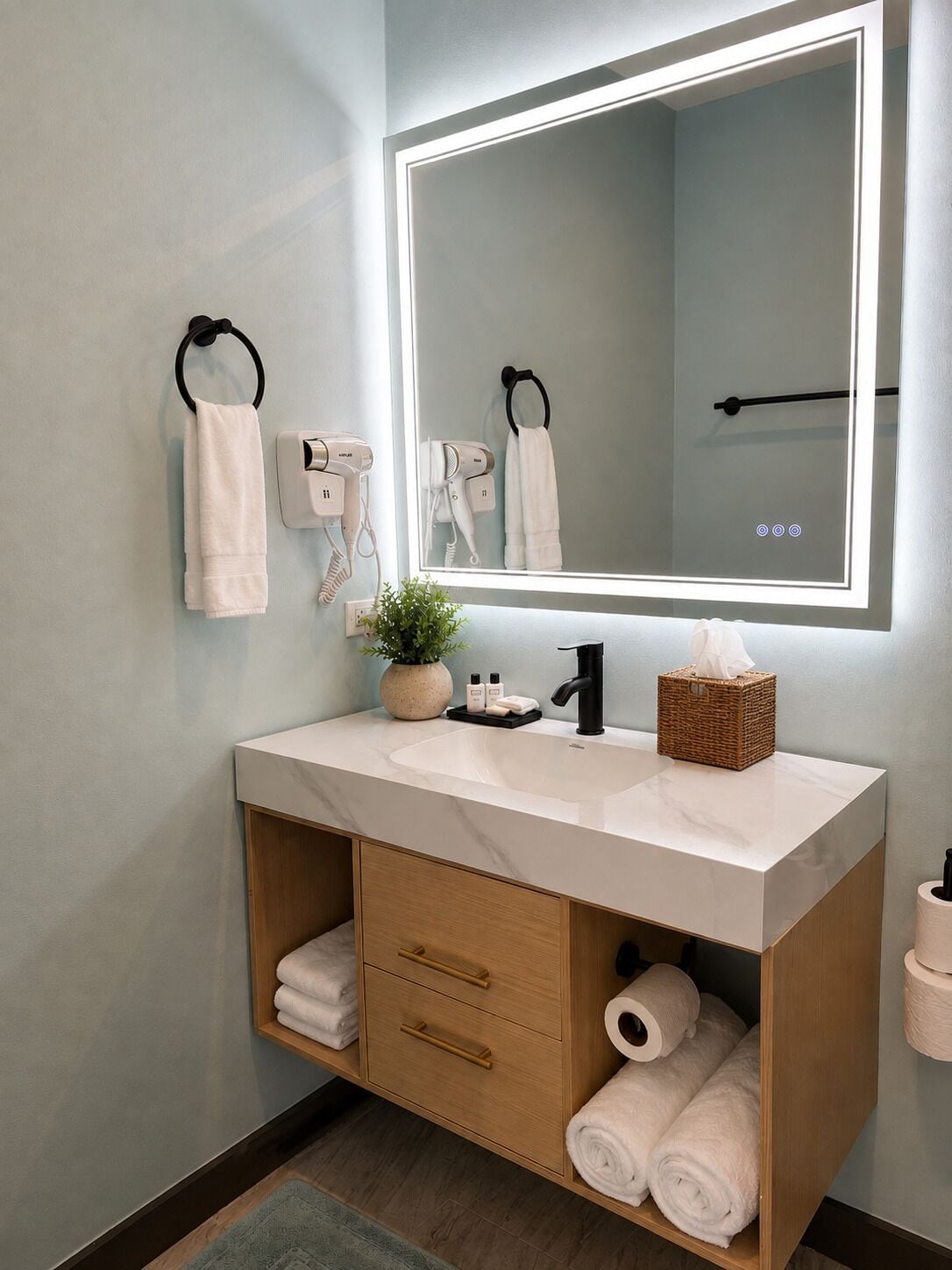 A bathroom with a large mirror, towel racks, a hairdryer, a sink, a plant, a tissue box, and a toilet paper holder.
