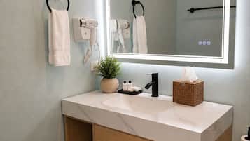 A bathroom with a large mirror, towel racks, a hairdryer, a sink, a plant, a tissue box, and a toilet paper holder.