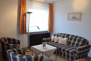 Ein Zimmer mit einem Karomuster-Sofa, zwei Sesseln, einem kleinen Tisch mit einem Glas, einer Stehlampe und einem Bild an der Wand.