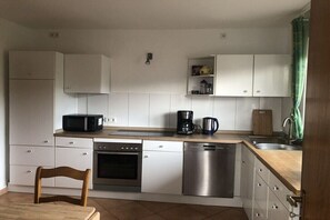 Eine kleine Küche mit weißen Schränken, einer Mikrowelle, einer Kaffeemaschine, einem Geschirrspüler und einem Holzbstuhl.