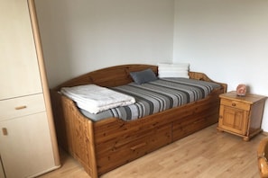 Ein hölzernes Bett mit einer gestreiften Tagesdecke, einem weißen Kopfkissen und einem grauen Kissen. Ein Holzbeistelltisch mit einer Vase darauf.