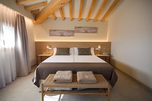 A hotel room with a bed, two bedside tables, a bench with towels, wooden beams on the ceiling, and two wall-mounted artworks.